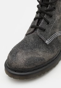 Svart gummisåle støvel i grå filtmateriale, med svarte snører, metalløye og kontrasterende hvite sømmer på overdelen.