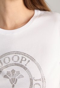 Camiseta de algodón blanca con escote redondo y adornos de brillantes plateados que forman texto y patrones circulares en la parte frontal.