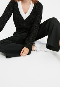 Pull scintillant noir avec un col en V blanc, associé à un pantalon noir à jambes larges et des baskets blanches. Mains posées sur le pantalon.