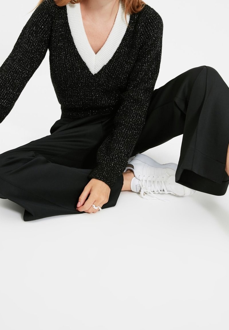 Pull scintillant noir avec un col en V blanc, associé à un pantalon noir à jambes larges et des baskets blanches. Mains posées sur le pantalon.