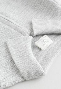Jasnoszary polarowy materiał o teksturowanym splotem, z prążkowanymi mankietami i małą metką oznaczoną "NEXT Baby". Miły w dotyku i przytulny wygląd.