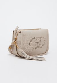 Sac bandoulière en cuir beige avec un design de logo perforé, fermeture zippée et accent en frange. Texture lisse et forme rectangulaire.