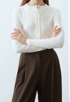 Persona che indossa una maglia bianca a maniche lunghe con bottoni, con le braccia incrociate e pantaloni largi a vita alta di colore marrone scuro, su uno sfondo neutro.