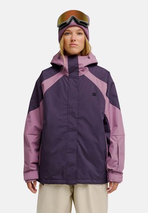 Femme portant une veste de ski violette et rose, un bonnet violet et des lunettes de ski sur le front, debout devant un fond uni.