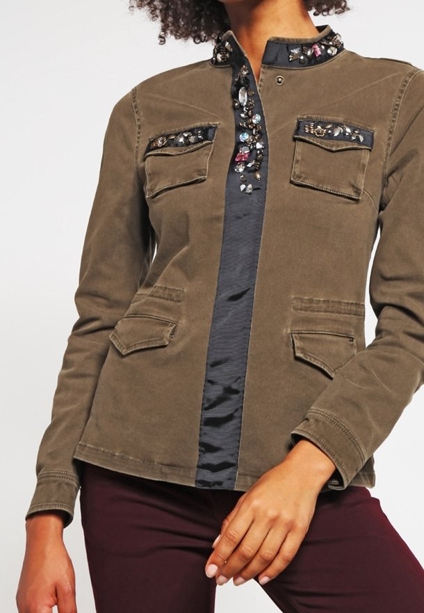 Femme portant une veste vert olive avec un col et des rabats de poche ornés de bijoux, associée à un pantalon violet foncé.