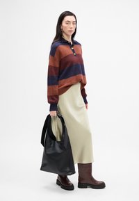 Pull en tricot rayé rouge, marron et bleu marine, assorti à une jupe crème et des bottes marron mi-mollet. Sac en cuir noir avec des anses longues.