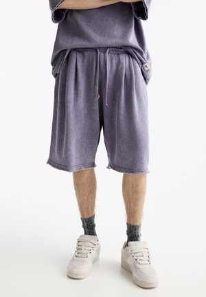 Person wearing loose purple shorts with drawstring, matching oversized top, gray socks, and white sneakers, standing with arms crossed.