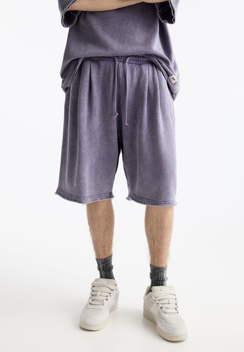 Personne portant un short violet ample avec un cordon de serrage, un haut oversize assorti, des chaussettes grises et des baskets blanches, debout les bras croisés.