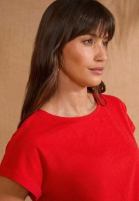 Top corto de manga corta rojo con cuello redondo, hecho de una tela suave. Presenta un detalle de puños enrollados y un brillo sutil. Diseño minimalista.