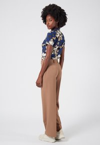 Bloemenprint shirt met blauwe achtergrond, korte mouwen; bruine wijde broek; witte sneakers; model poseert met de rug naar de camera.