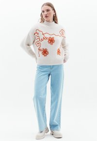 Maglione lavorato a maglia color crema con ricami floreali arancioni, colletto alto e orlo a coste. Abbinato a jeans a palazzo azzurri e scarpe beige.