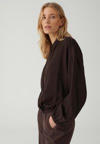 Donna bionda che indossa una giacca marrone scuro larga con bottoni e polsini elasticizzati e pantaloni abbinati, in piedi con una mano in tasca.