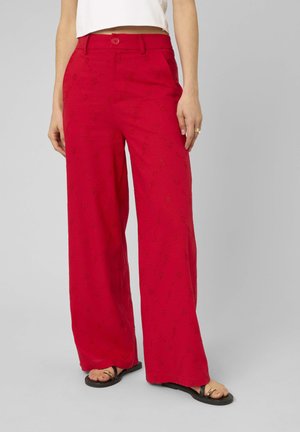 Femme portant un pantalon rouge taille haute à jambes larges avec une broderie florale subtile et des sandales noires sur fond gris.