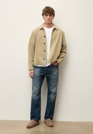 Ung mand stående iført beige knappet jakke, hvid t-shirt, blå jeans og lysebrune loafers mod en simpel lys baggrund.