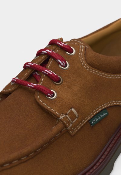 Καφέ παπούτσι από σουέντ με κόκκινα και λευκά κορδόνια, λευκή ραφή και μια μικρή πράσινη υφασμάτινη ετικέτα PS Paul Smith στο πλάι.