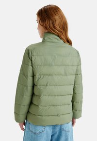 Ljusgrön puffjacka med horisontell quiltning, hög krage och långa ärmar. Återgiven med blå denim, sedd bakifrån.