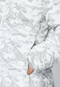 Veste matelassée gris clair avec un motif de montagne blanc, dotée d'une poche zippée et de coutures apparentes. Texture de tissu douce.