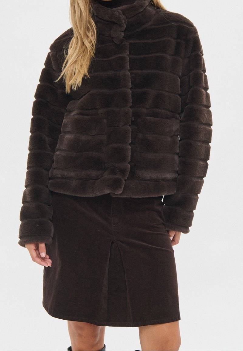 Femme portant une veste en fourrure pelucheuse marron foncé et une jupe assortie longueur genou avec une fente à l'avant, debout devant un fond clair.