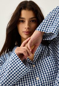 Niebieska i biała koszula w kratkę z kołnierzykiem, dużymi mankietami i guzikami, prezentująca bawełnianą teksturę i luźny krój.