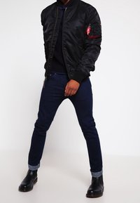 Schwarze Nylon-Bomberjacke mit Reißverschluss, gerippten Bündchen und einer Seitentasche mit rotem Akzent. Kombiniert mit dunkelblauen Jeans und schwarzen Stiefeln.