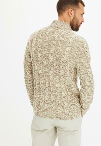 Maglione beige a maglia con motivi testurizzati, colletto ripiegato e orlo a costine. Indossato sopra pantaloni chiari.