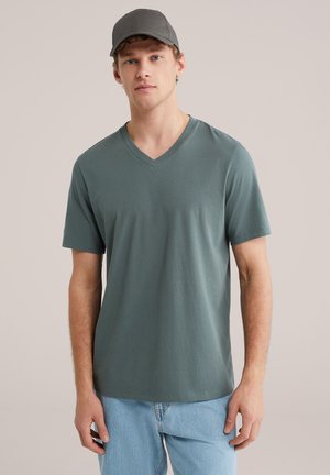 Jonge man met een grijze baseballpet, groene V-hals T-shirt en lichtblauwe spijkerbroek, staand tegen een effen achtergrond.