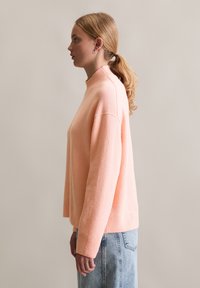 Maglione a maglia rosa chiaro con collo alto e polsini a costine, abbinato a jeans blu a vita alta. Texture morbida, vestibilità comoda e design senza cuciture.