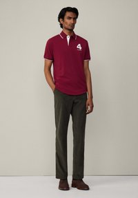 Burgundská polo košeľa s bielymi akcentmi a logom čísla "4", skombinovaná s tmavozelenými nohavicami a hnedými topánkami. Hladká tkanina, klasický strih.