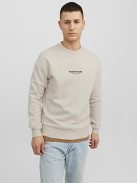 Beige sweatshirt met ronde hals, ribbelmanchetten en onderrand. Bevat het "Originals Studio" logo in zwart op de borst. Draagt bovenop lichte denim jeans.