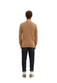 Mann mit lockigem Haar, der einen braun gemusterten Pullover, dunkle Jeans mit umgeschlagenen Säumen, weiße Socken und braune und weiße Turnschuhe trägt und mit dem Rücken zur Kamera steht.