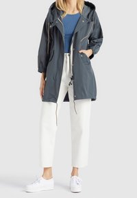 Czarnoszara kurtka przeciwdeszczowa z kapturem, elastycznymi mankietami i ściąganym dołem, noszona z niebieską bluzką, białymi spodniami i białymi trampkami.