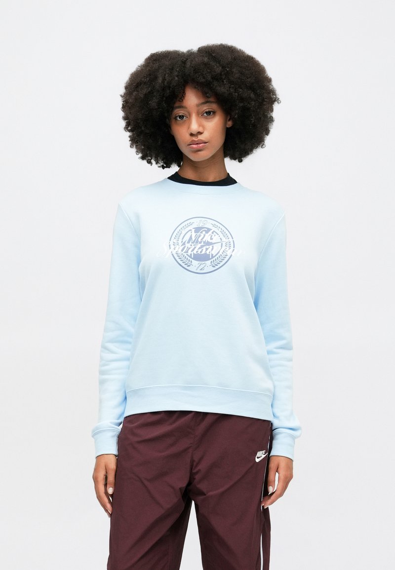 Sweat-shirt bleu clair avec un logo blanc rond au centre, des poignets côtelés et un tissu texturé, porté avec un pantalon marron foncé.