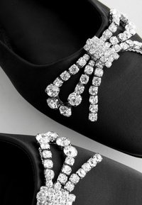 Musta satiininen ballerina-kengät, joissa on suuri strassikristallista tehty nauha-akkentti. Nauhassa on keskellä kristalliryhmä, jossa useat kirkkaat kivet on aseteltu symmetrisesti.