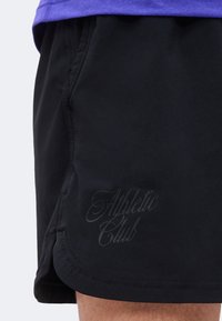 Μαύρα αθλητικά σορτς με διακριτικό κείμενο "Athletic Club" στο αριστερό μηρό, που φοριούνται από άτομο με ορατό πόδι και μωβ τελείωμα στο πουκάμισο.