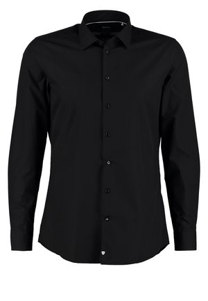 Mann trägt schwarzes Hemd mit Knöpfen und marineblaue Hose, hält schwarze Reisetasche, geht mit einem Fuß nach vorne vor einfarbigem Hintergrund.