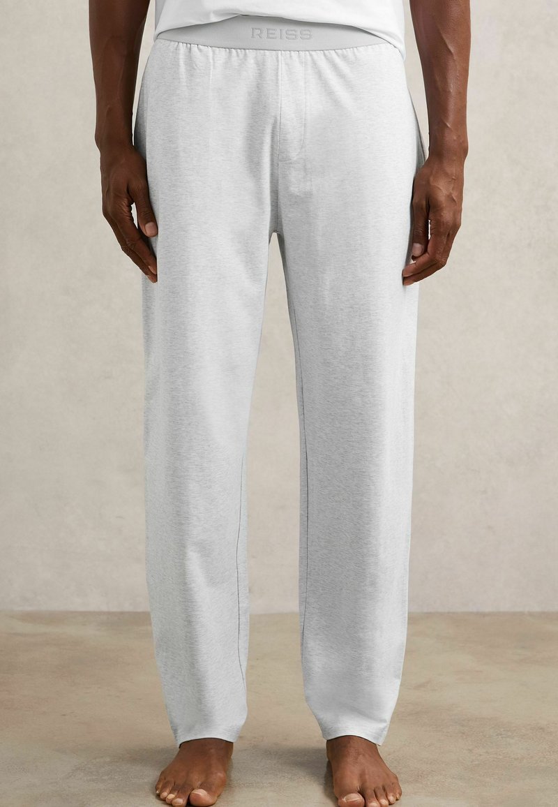 Uomo che indossa pantaloni della tuta grigio chiaro e una camicia bianca, in piedi a piedi nudi su un pavimento neutro davanti a uno sfondo semplice.