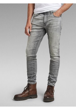 Homme portant un jean slim gris délavé et des bottines en cuir marron foncé à lacets, main dans la poche, debout sur un fond gris.