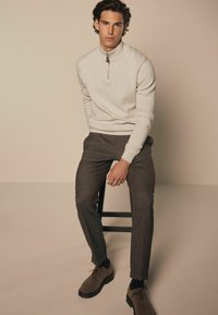 Lys beige zip-sweater med høj krave, parret med mørkebrune skræddersyede bukser og brune sko, siddende på en sort skammel mod en neutral baggrund.