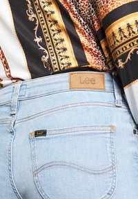 Jeans de mezclilla azul claro con costuras naranjas, que presentan una etiqueta de cuero con la inscripción "Lee" en la parte trasera de la cinturilla y una pequeña etiqueta negra en el bolsillo.