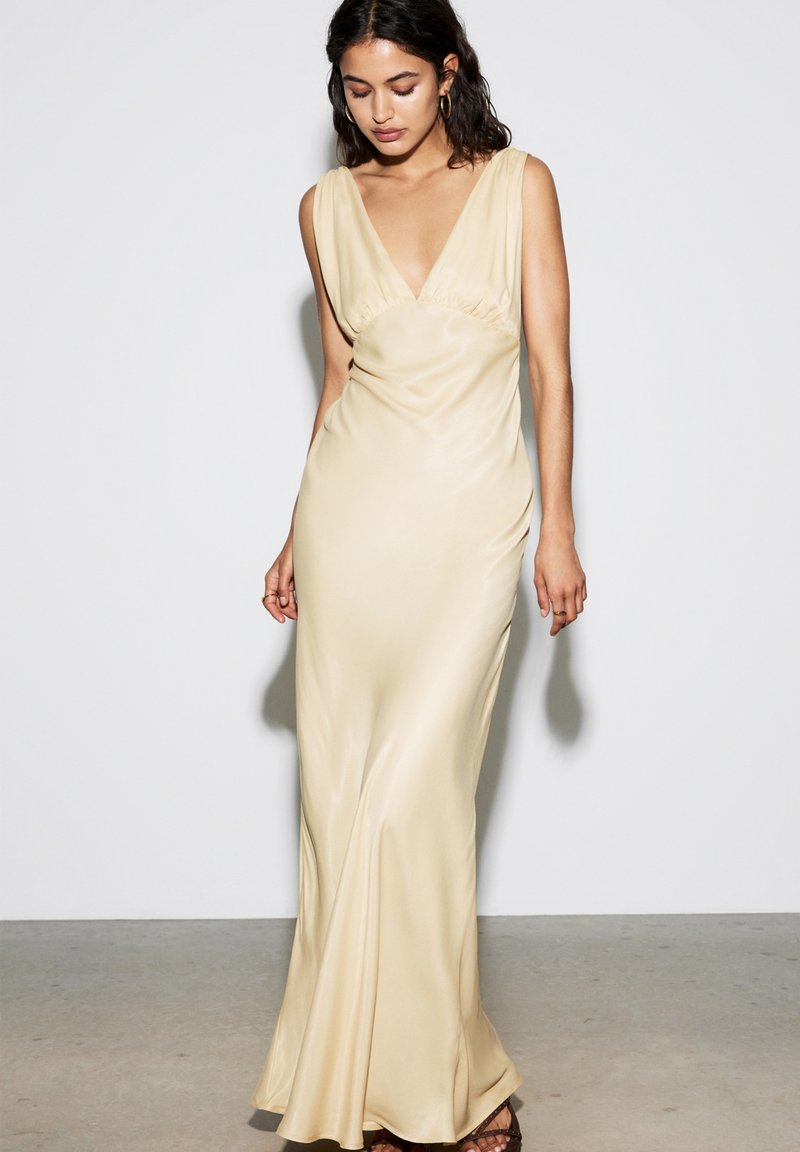 Robe de soirée cylindrique en satin beige clair, dotée d'un décolleté en V profond, sans manches, avec un drapée délicate qui cascade jusqu'au sol.