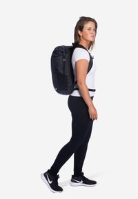 Zwarte rugzak met een gestructureerde oppervlakte, verstelbare banden en een heupriem, gedragen door een persoon in een strakke witte blouse en zwarte leggings.