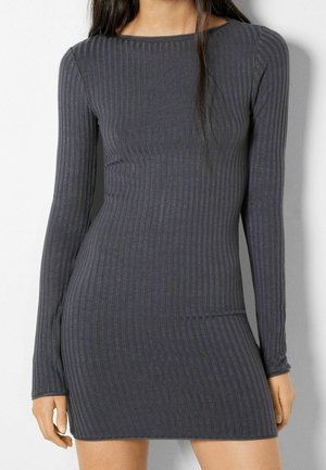 Fitted ribbed long-sleeve dark gray mini dress on person standing against white wall, showing torso and upper legs.