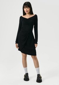 Zwarte jurk met lange mouwen en een sweetheart-halslijn, gerimpelde buste, knoopdetail bij de taille en een gedrapeerde rok.