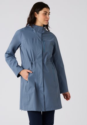 Femme portant un imperméable bleu mi-genoux avec capuche, poches zippées sur la poitrine et les côtés, debout avec une main dans la poche.