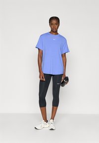 Μπλε κοντομάνικο T-shirt της Nike συνδυασμένο με μαύρο κολάν κάπρι. Το μοντέλο κρατάει ένα μαύρο μπουκάλι νερού, με ορατά λευκά αθλητικά παπούτσια.