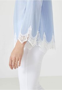 Ljusblå blus med intrikat vitt spetsdetalj vid ärmarna och nederkanten, gjord av ett mjukt tyg. Bärs med vita byxor.