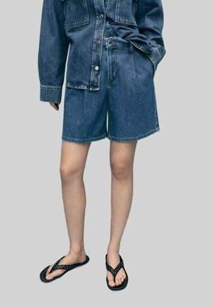 Denim shorts in dark blue, featuring a relaxed fit and rolled hem. Paired with black flip-flops adorned with silver studs.