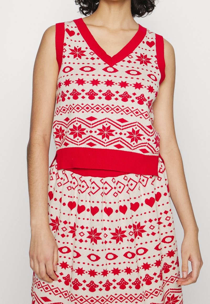 Femme portant un gilet sans manches en tricot rouge et blanc et une jupe assortie avec des motifs de cœur, étoile, œil, champignon et géométriques.