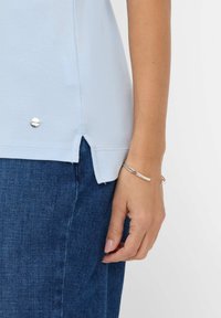 Lysblå polo t-shirt med en tekstureret finish, der har sideslids og en sølvlogo aksent ved kanten. Kombineret med mørke denim.