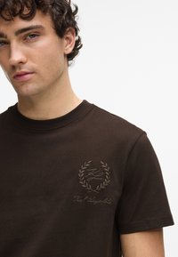Brūna kokvilnas T-krekls ar apaļu kaklu, ar strukturētu logo un lauru dizainu izšūtu kreisajā krūtī.